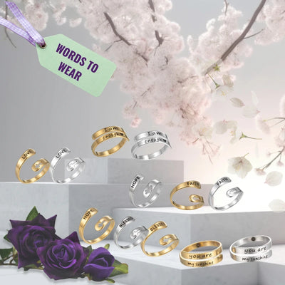 Collection of gold and silver rings with validation messages laser etched on, shown on a light gray background with cherry blossoms and purple roses.