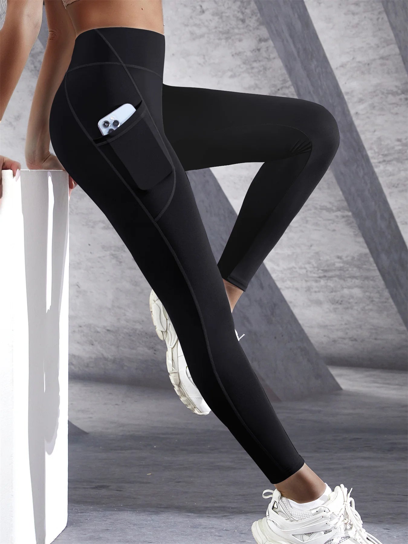 Woman wearing black high waist leggings with a side pocket and phone inside the pocket leaning on bench with a light background