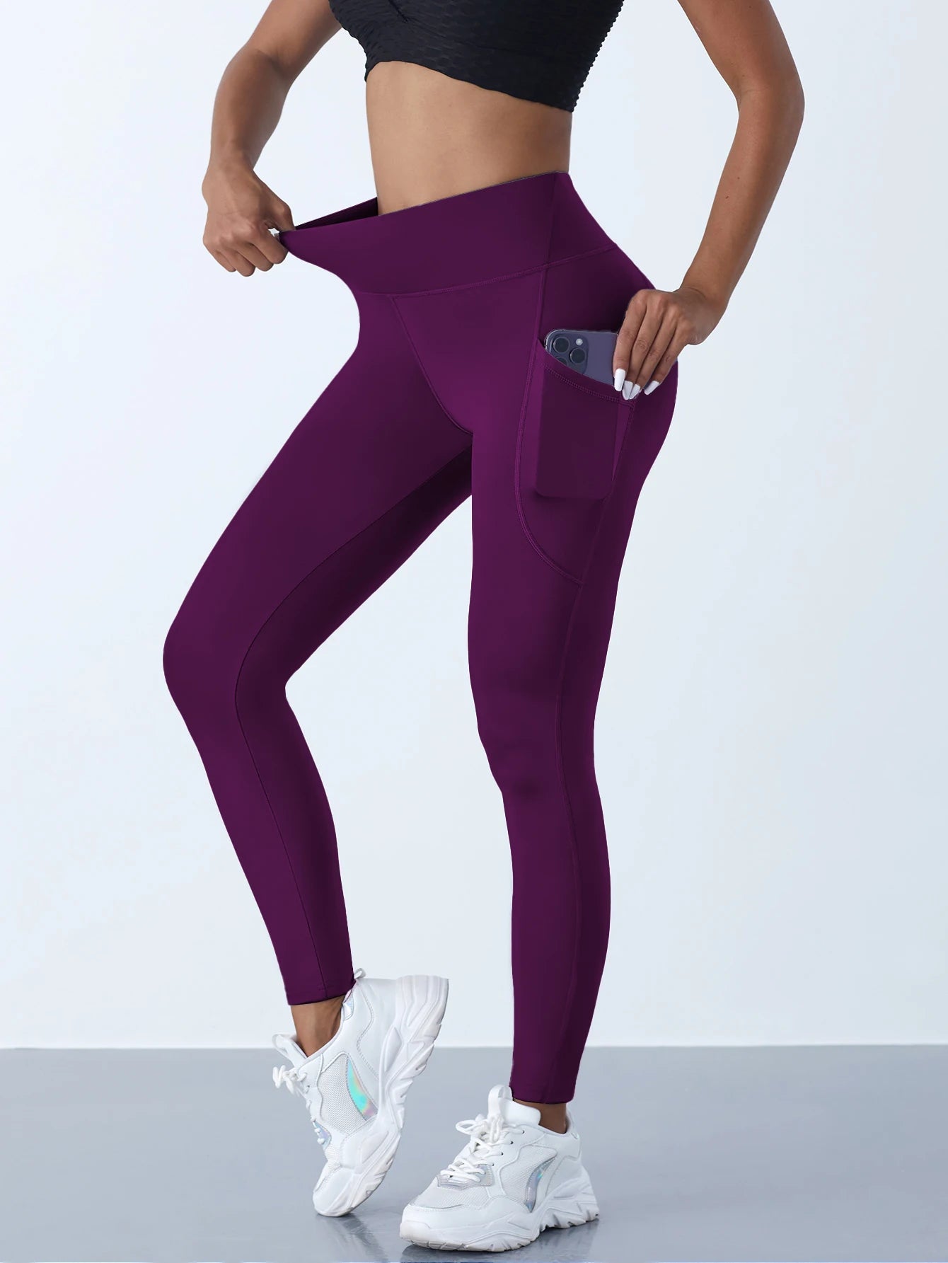 Woman wearing dark purple high waist leggings with a side pocket and phone inside the pocket showing stretchable fabric with a light background