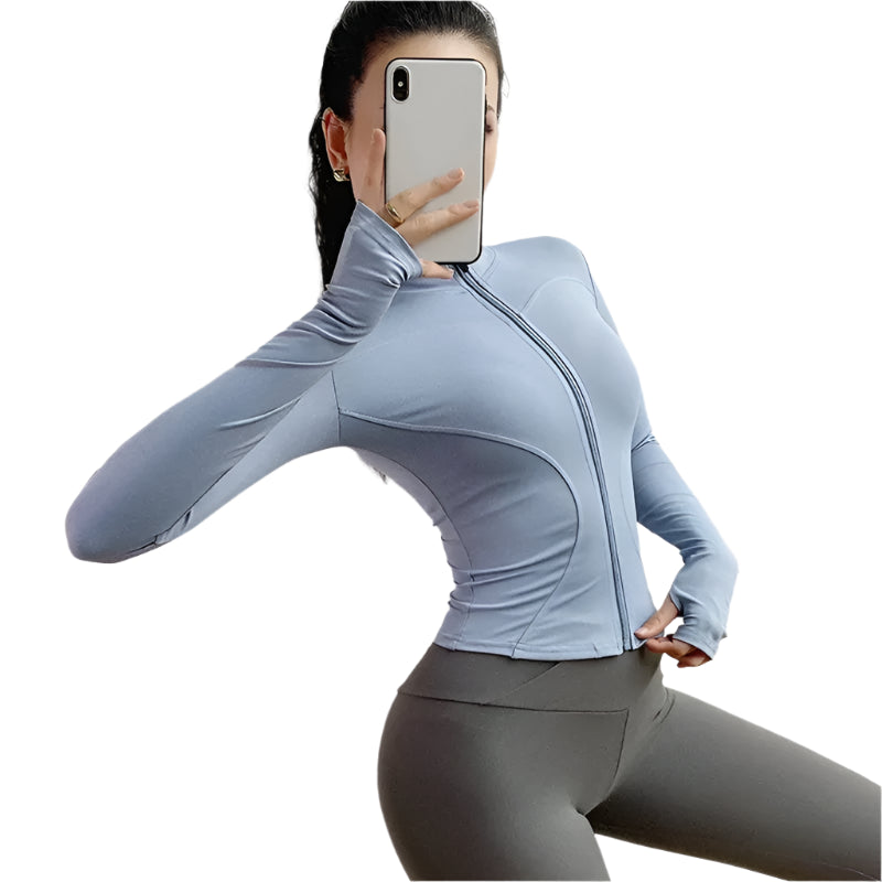 Woman wearing a light blue zip up thumb hole jacket and gray leggings taking a selfie against a white background 