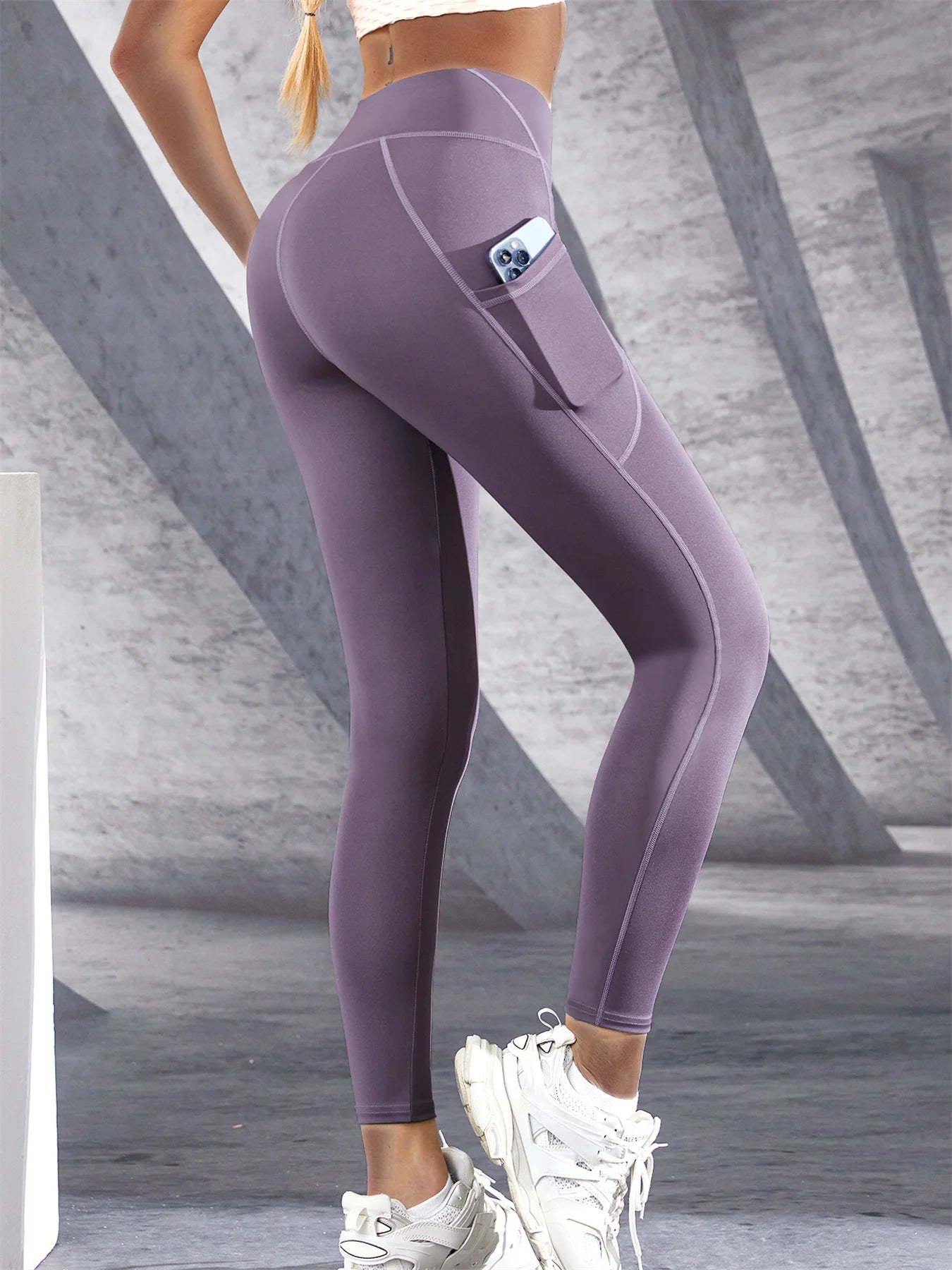 Woman wearing purple high waist leggings with a side pocket and phone inside the pocket on a light background