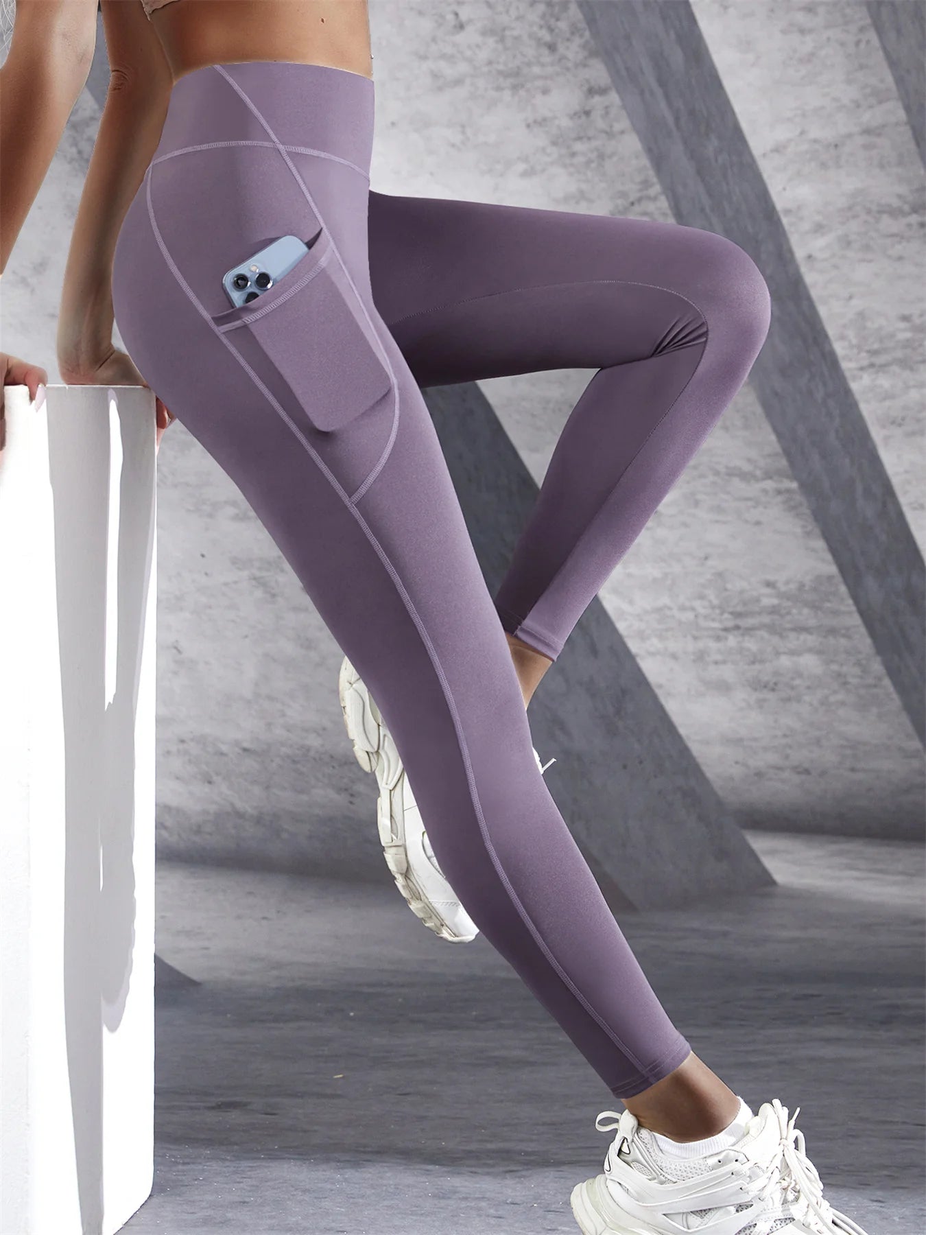 Woman wearing purple high waist leggings with a side pocket and phone inside the pocket leaning against bench on a light background